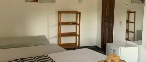 Un dormitorio con cama, mesita de noche con un sombrero, una silla, estante de madera, aire acondicionado blanco y un cuadro en la pared.