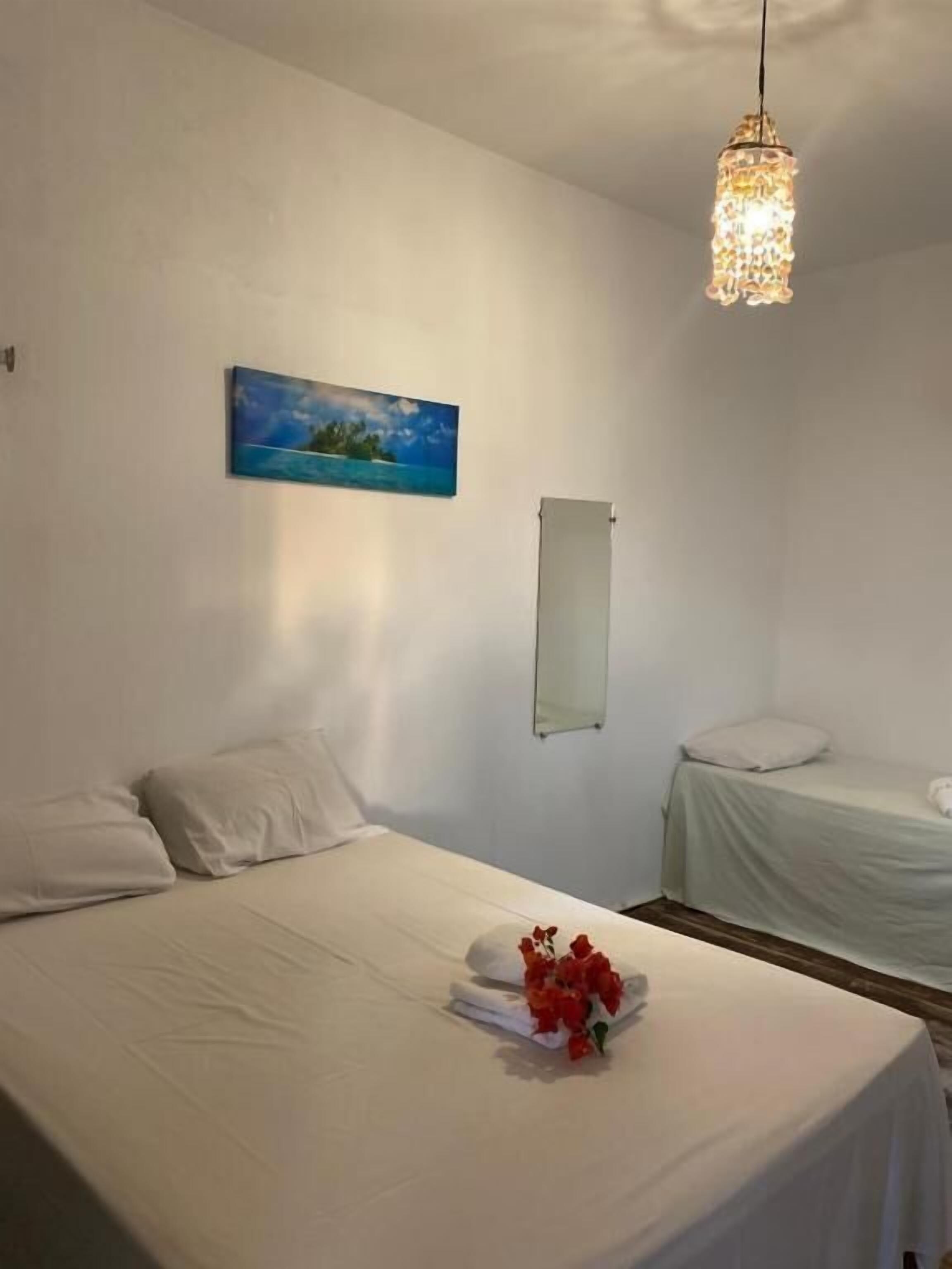 A hotel room with a bed, a bedside table with a towel and flowers, a hanging light fixture, a painting on the wall, and a small window.