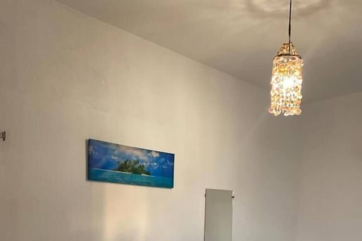 A hotel room with a bed, a bedside table with a towel and flowers, a hanging light fixture, a painting on the wall, and a small window.