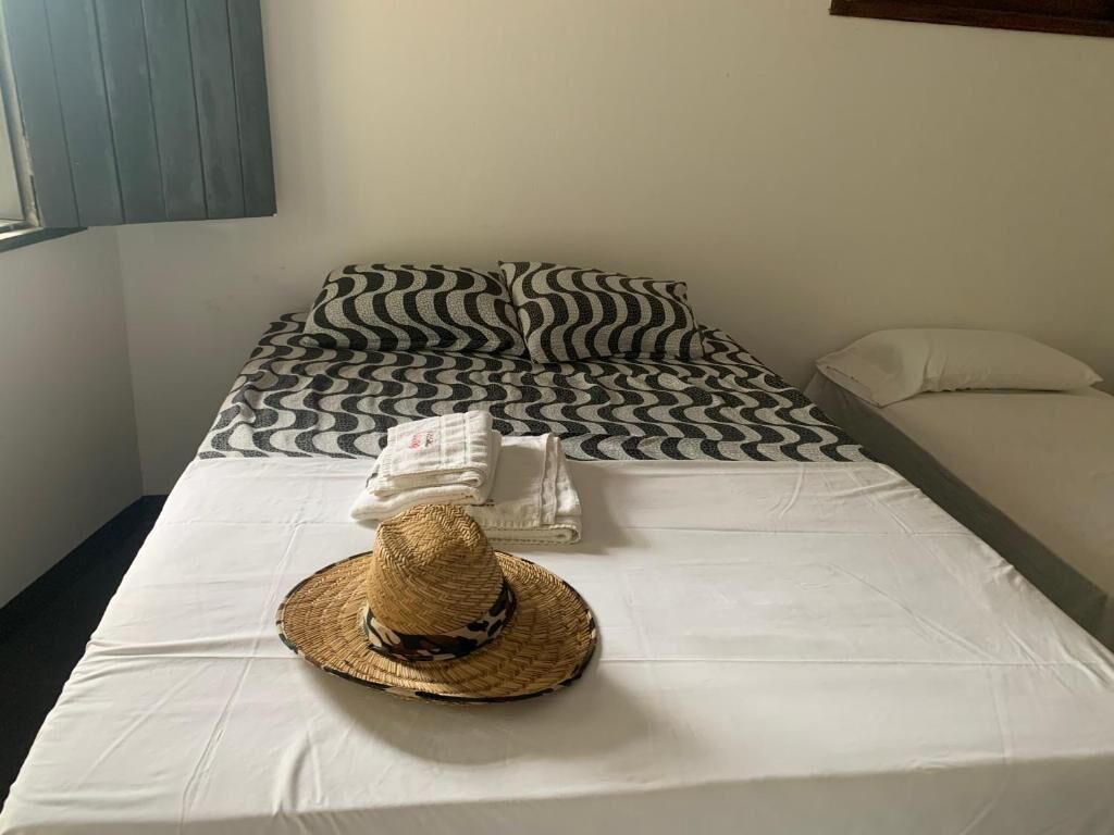 Una habitación de hotel con una cama cubierta por una colcha estampada en blanco y negro, un almohadón blanco y una manta blanca. Un sombrero de paja está sobre la cama.