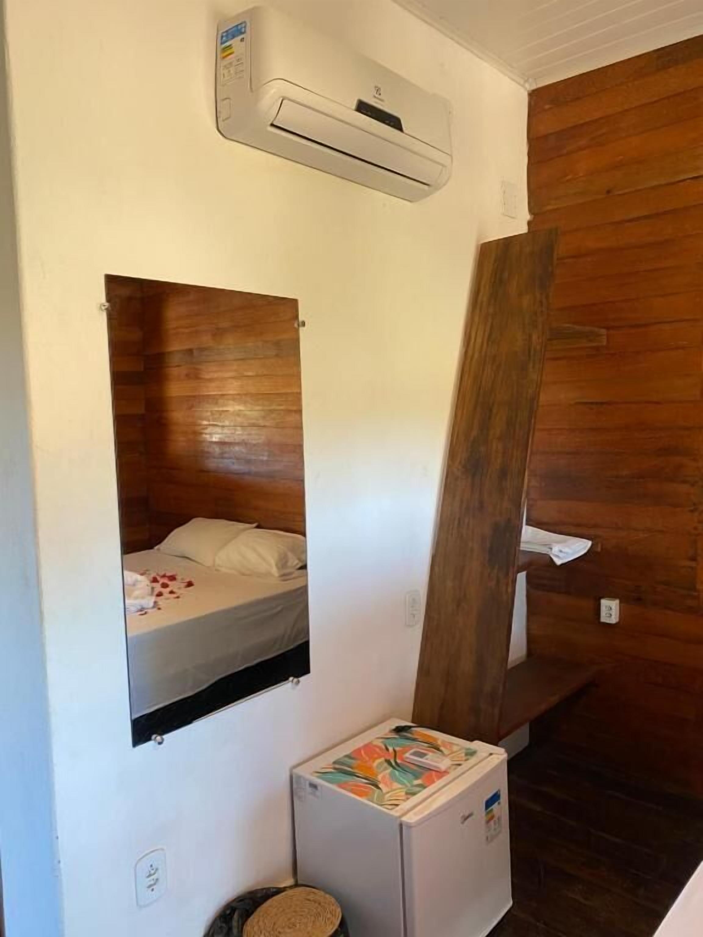 A compact room with a bed, a small refrigerator, a mirror, and an air conditioning unit.