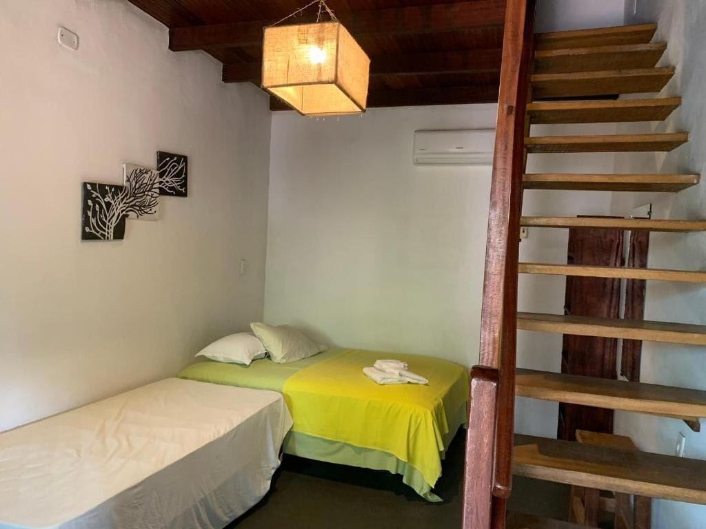 Una habitación con dos camas, una lámpara colgante, un equipo de aire acondicionado y una escalera de madera.
