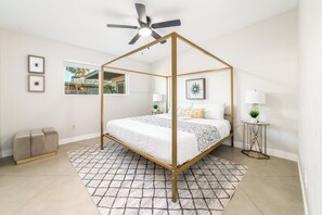 Un lit à baldaquin à quatre poteaux avec un ventilateur de plafond, une petite table de chevet avec une lampe et une vue sur l’extérieur par une fenêtre.