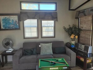 Ein Wohnzimmer mit einer Couch, einem Billardtisch, einem Ventilator und einem Fenster mit Vorhängen.