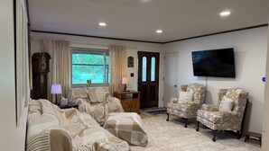 A living room with a sofa set, a television, a clock, and a window with curtains. - 4BR Home - Airport- UD - WPAFB - POOL -Sleeps 7 (Dayton)