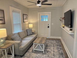 A living room with a sofa, a coffee table, a lamp, a ceiling fan, a door, a TV, and a patterned rug.