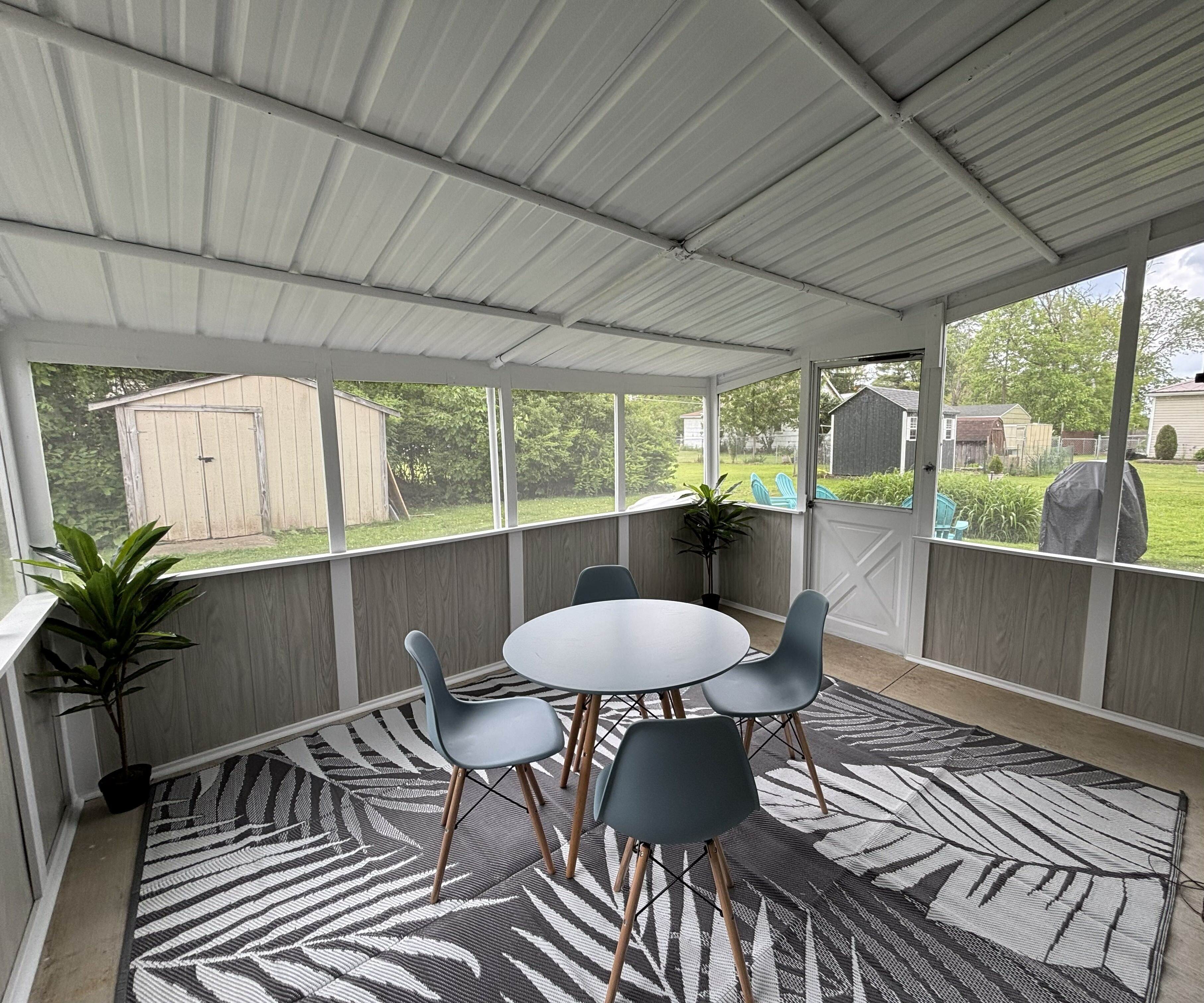 A covered patio with a round table and four chairs, a patterned rug, and a view of a backyard.