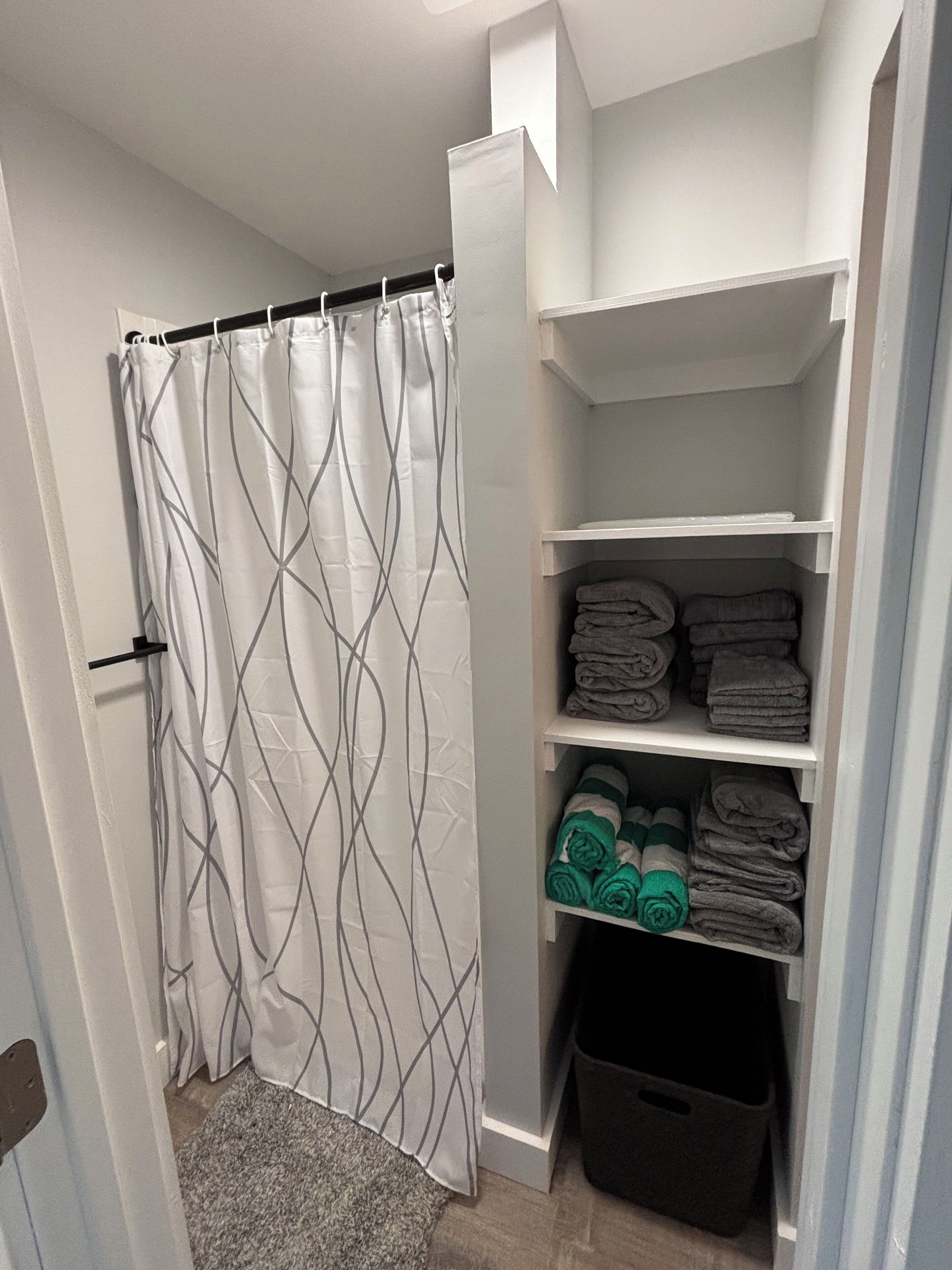Un baño con cortina de ducha, toallero y estante con toallas dobladas y una cesta de ropa.