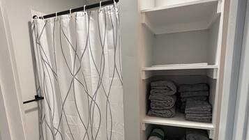 Un baño con cortina de ducha, toallero y estante con toallas dobladas y una cesta de ropa.