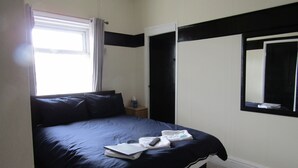 Quarto com cama, mesa de cabeceira, janela com cortinas, espelho e porta.