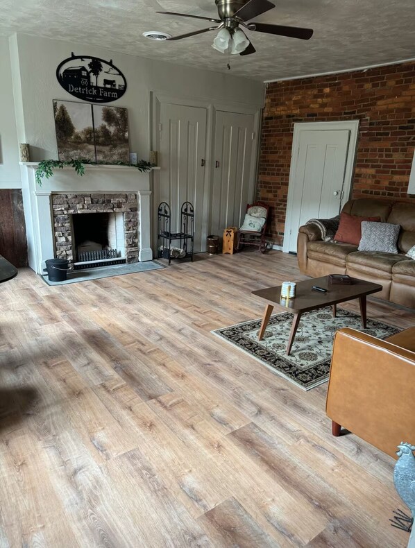 A living room with a fireplace, a ceiling fan, a brick wall, a sofa, and a coffee table. - 3-bedroom farmhouse in tranquil Bedford with cozy charm (Bedford)