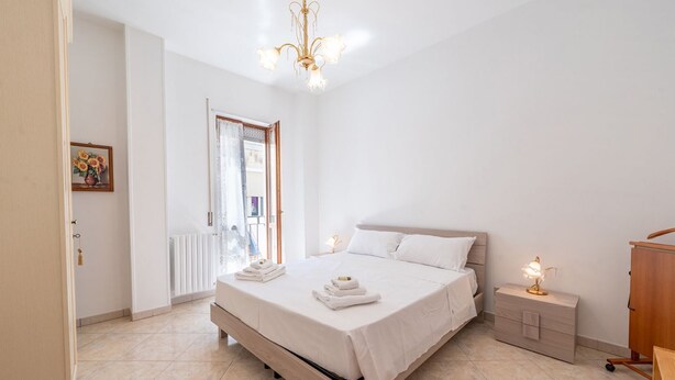 A bedroom with a bed, bedside tables, a nightstand, a dresser, a chandelier, and a door leading to a balcony. - Nacci White Home by Rentbeat (Ostuni)
