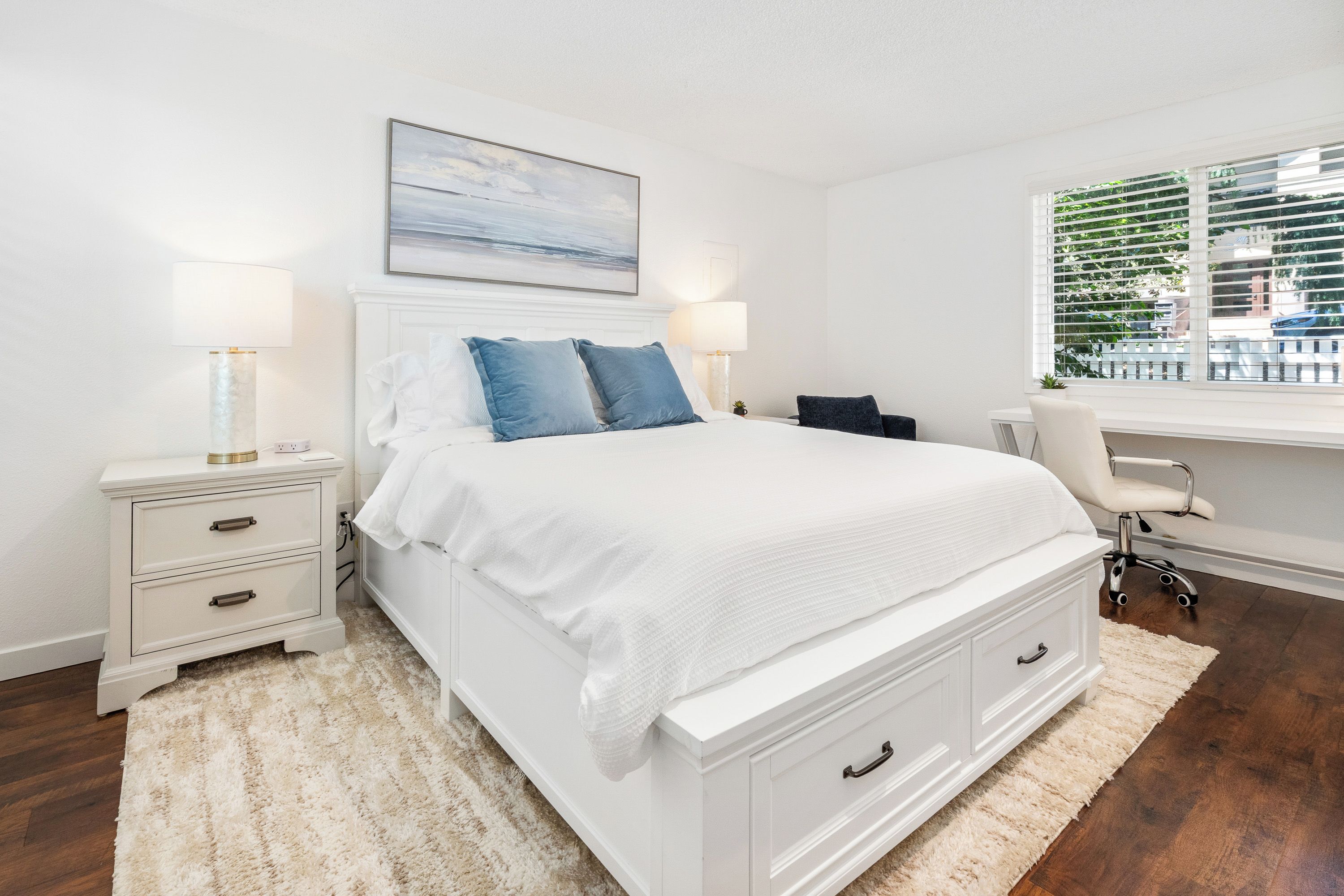 A bedroom with a white bed, two bedside tables, a desk, a chair, a window with blinds, and a painting on the wall.