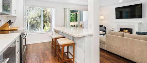 A modern kitchen with a central island, bar stools, and a dining area with a view of greenery.