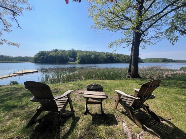Eine Uferlandschaft mit zwei Adirondack-StĂŒhlen und einem kleinen Tisch, umgeben von einer RasenflĂ€che und einem groĂen Baum.