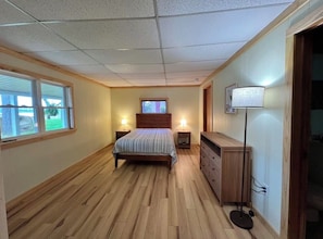 Quarto com piso de madeira, uma cama com colcha listrada, duas mesas de cabeceira com abajures, um penteadeira e uma janela com vista para o verde.