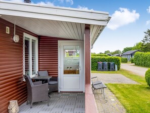 Ett takförsett utomhusområde med ett utebord och stolar, en dörr med ett glasparti och en gångväg som leder till en rad sopkärl.