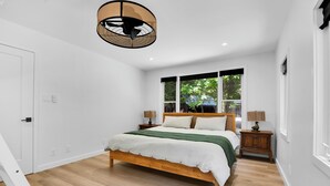 Un dormitorio con cama de madera, paredes blancas, ventana con vistas a zonas verdes y ventilador de techo.