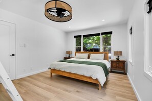 A bedroom with a wooden bed, white walls, a window with a view of greenery, and a ceiling fan.