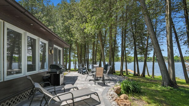 Un patio con mobiliario exterior, una parrilla y vistas a un lago rodeado de árboles.
