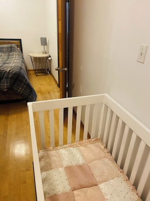 Quarto com berço, cama com colcha xadrez, mesinha redonda com abajur e uma porta.