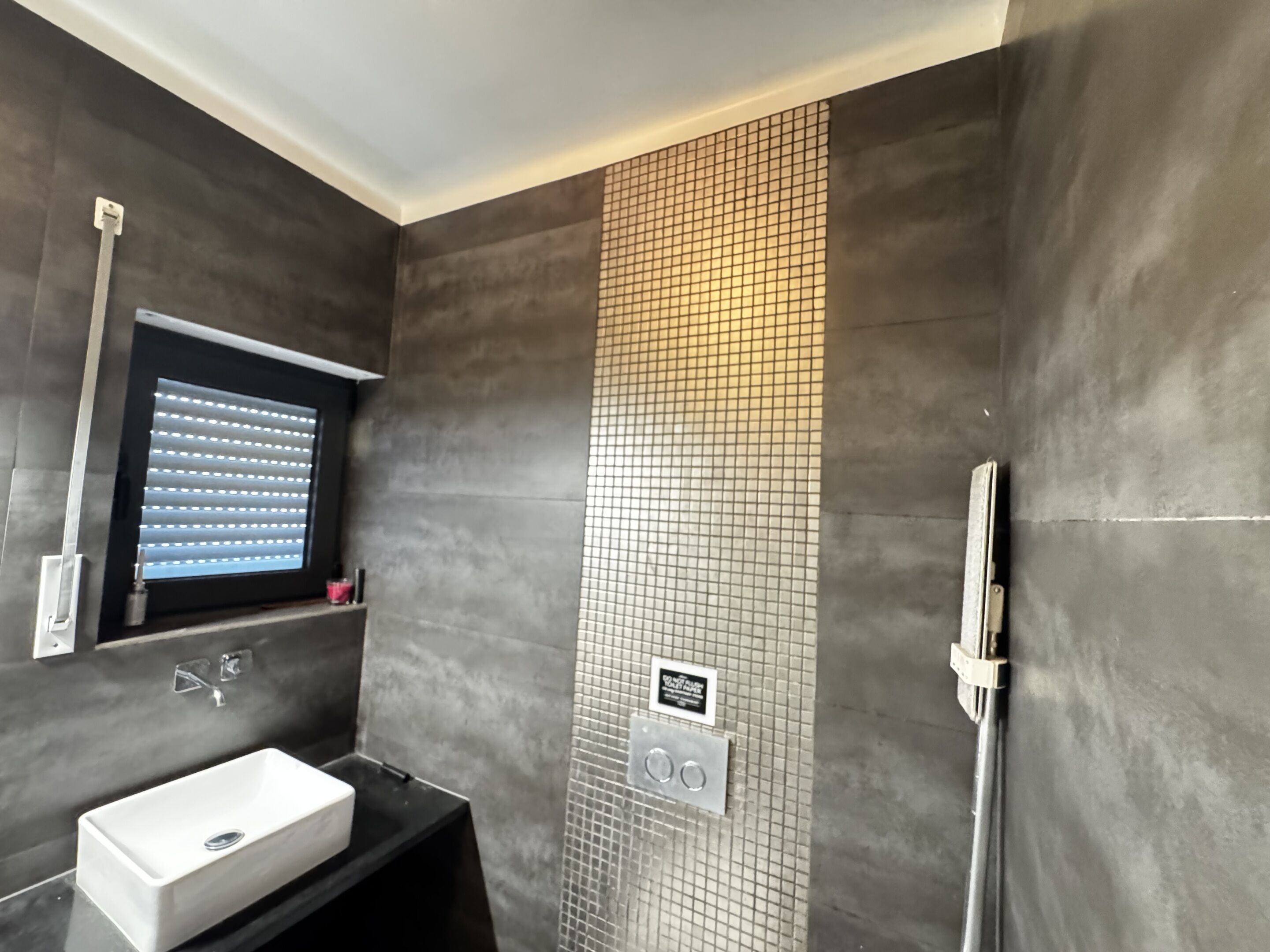 A modern bathroom with a rectangular sink, a window with blinds, a tiled shower wall, and a towel rack.