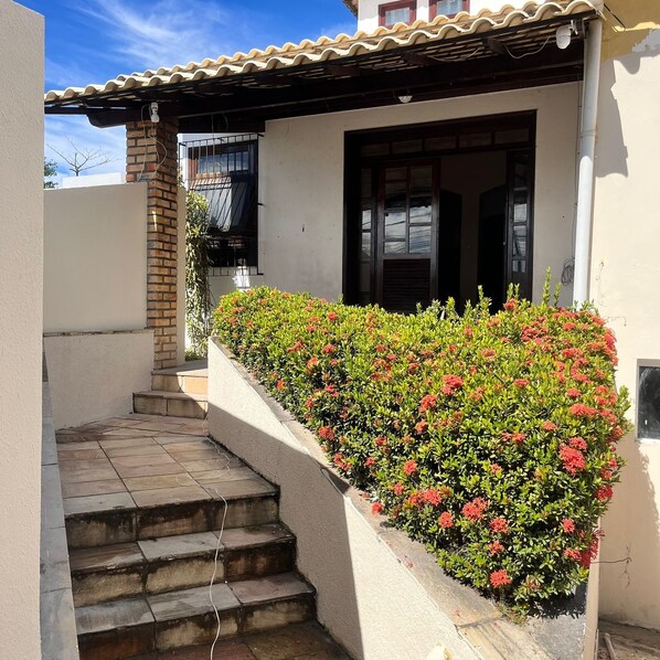 Une maison avec un toit en tuiles, une cheminée en briques, un escalier en béton et un parterre de fleurs rouges.