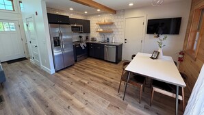 Cozinha moderna com eletrodomésticos de aço inoxidável, área de jantar com mesa e cadeiras, e TV fixada na parede.
