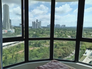 Quarto com uma janela ampla que oferece vista para um horizonte urbano com edifícios altos e áreas verdes.