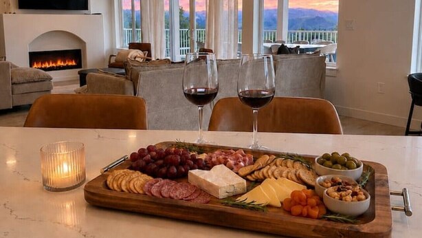 Un salon avec une cheminée, une table à manger agrémentée d’un plateau de charcuterie et un balcon offrant une vue sur le coucher de soleil.