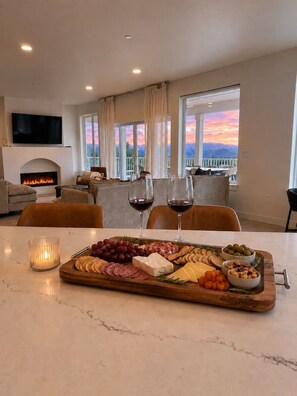 A living room with a fireplace, a dining table with a charcuterie board, and a balcony with a sunset view. - Pickleball - Fire Pit - EV Charger - Swim Spa (East Wenatchee)