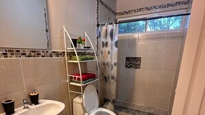 Une salle de bain avec un lavabo, des toilettes et une douche, le tout de couleur blanche. On y trouve un porte-serviettes avec des serviettes pliées et une plante en pot. La douche est dotée d’un rideau à motifs.