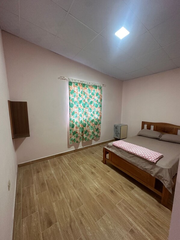 Ein Schlafzimmer mit einem Bett, einem Nachttisch, einem Wandregal und einem Fenster mit floralem Vorhang.