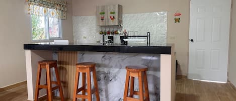 Una cocina con una isla de mármol, taburetes de madera, una puerta blanca y una ventana con cortinas.