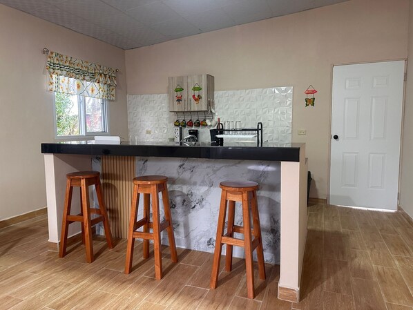 Une cuisine avec un îlot en marbre, des tabourets en bois, une porte blanche et une fenêtre avec des rideaux.