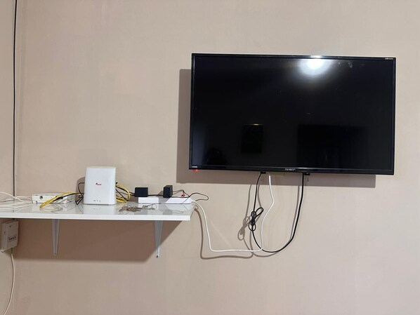 Un téléviseur à écran plat fixé au mur, avec une étagère blanche en dessous contenant des appareils électroniques et des câbles.