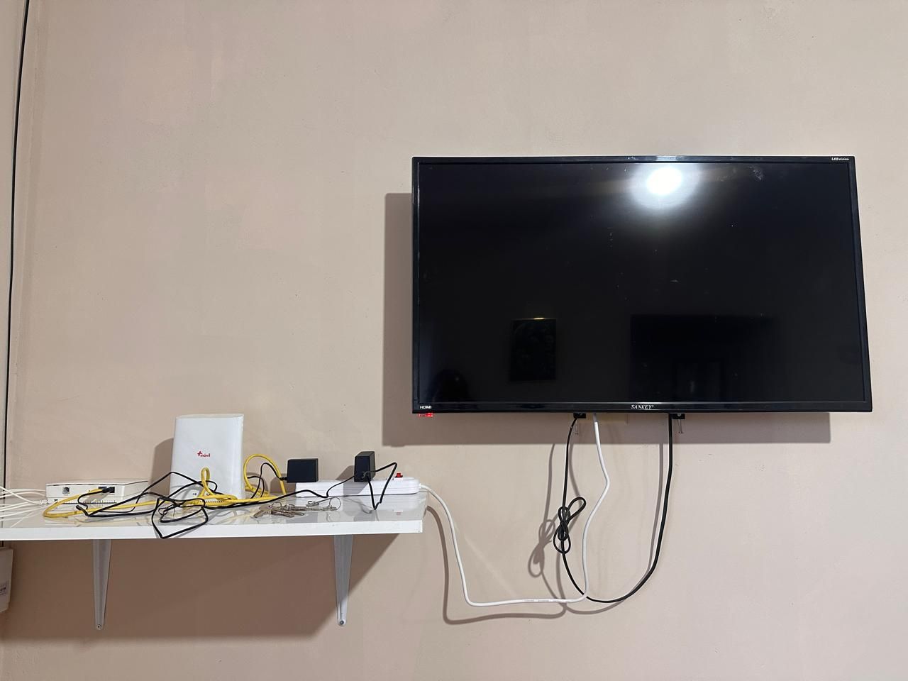 A flat-screen TV mounted on a wall with a white stand below it, connected to various electronic devices and cables.