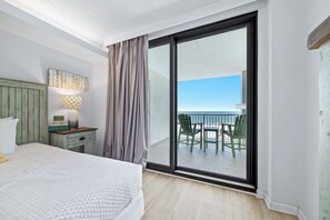 A bedroom with a bed, a nightstand, a lamp, a window with curtains, and a view of a balcony with chairs. - Summer House 404A | Oceanfront | Pools, Hot Tub, Tennis | My Beach Getaways (Orange Beach)