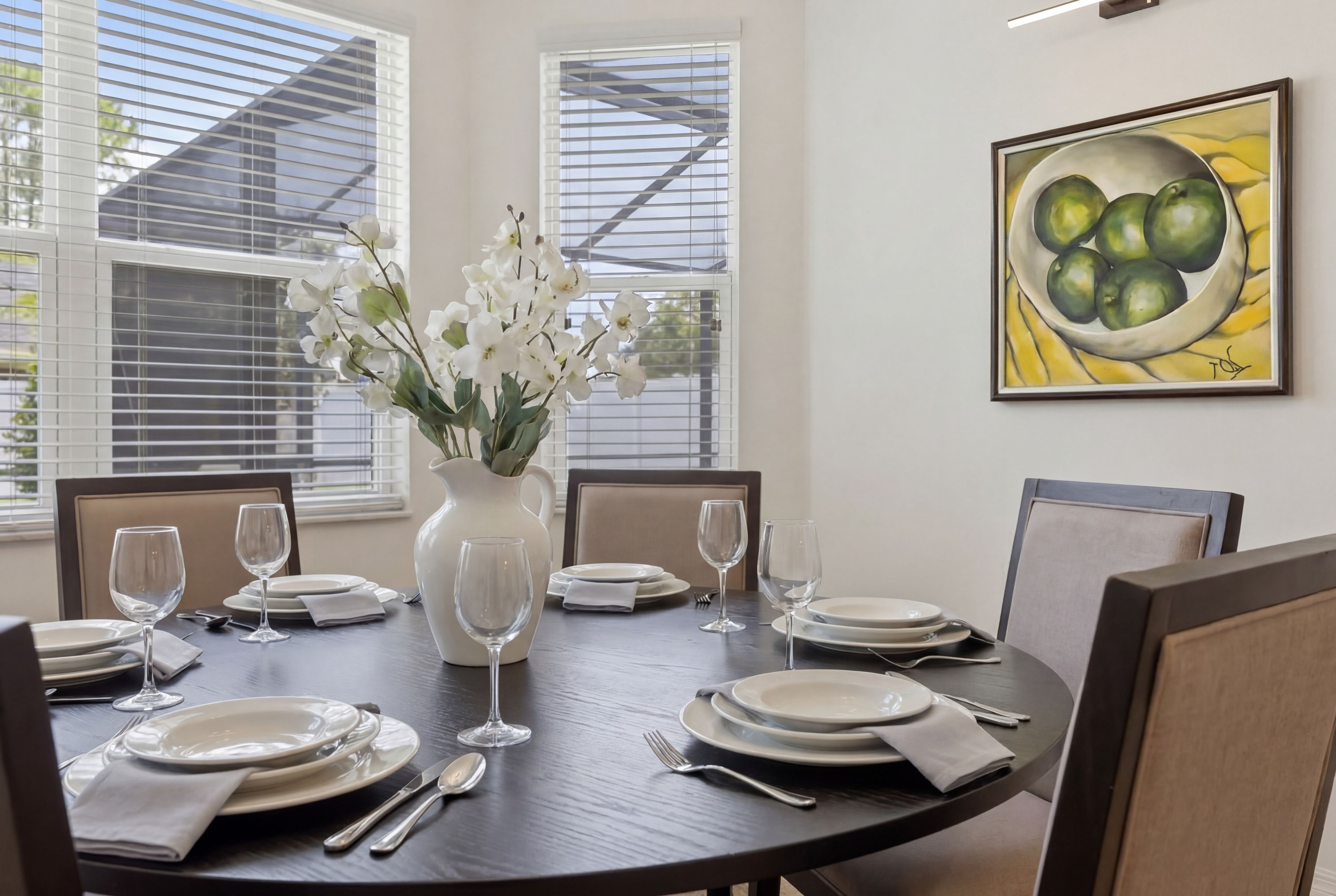 Mesa de comedor dispuesta para seis personas, con platos blancos, cubiertos y copas de vino transparentes. En el centro, un jarrón con flores blancas.