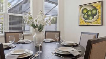 Mesa de comedor dispuesta para seis personas, con platos blancos, cubiertos y copas de vino transparentes. En el centro, un jarrón con flores blancas.
