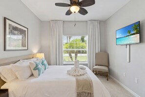Une chambre à coucher avec un lit, un ventilateur de plafond, une télévision, une chaise et une fenêtre avec des rideaux.