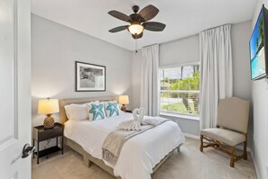 Une chambre à coucher comprenant un lit, une table de chevet, une chaise, un ventilateur de plafond, une fenêtre avec des rideaux et un téléviseur fixé au mur.