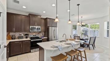 Una cocina moderna con armarios de madera oscura, electrodomésticos de acero inoxidable, una isla central con taburetes, iluminación colgante y vistas al exterior.