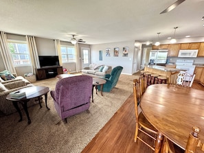 A living room with a sofa, a purple armchair, a coffee table, a TV, and a kitchen area with a dining table and chairs.