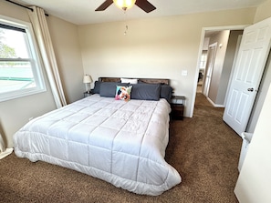 A bedroom with a large bed, a ceiling fan, a nightstand, a lamp, and a door leading to another room.