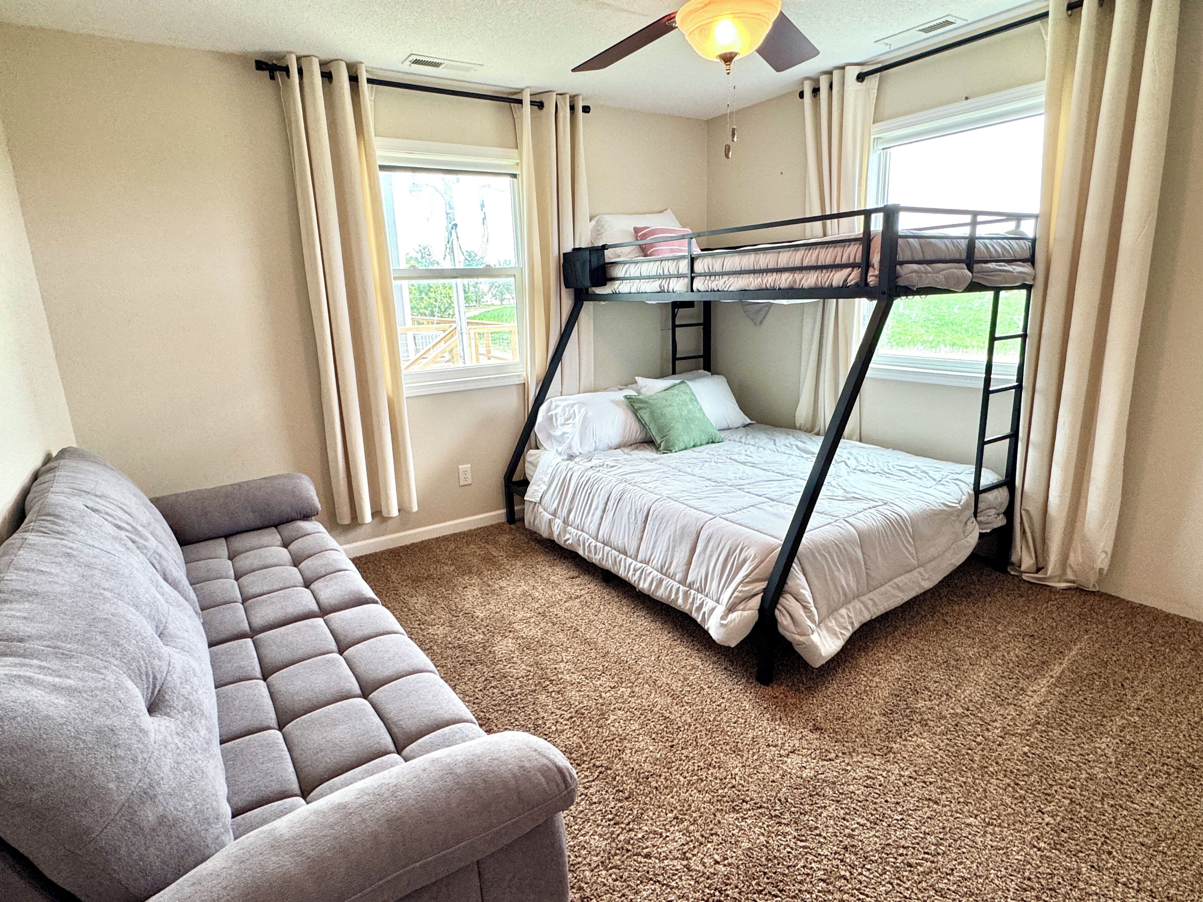 A room with a bunk bed, a sofa, a ceiling fan, and windows with curtains.