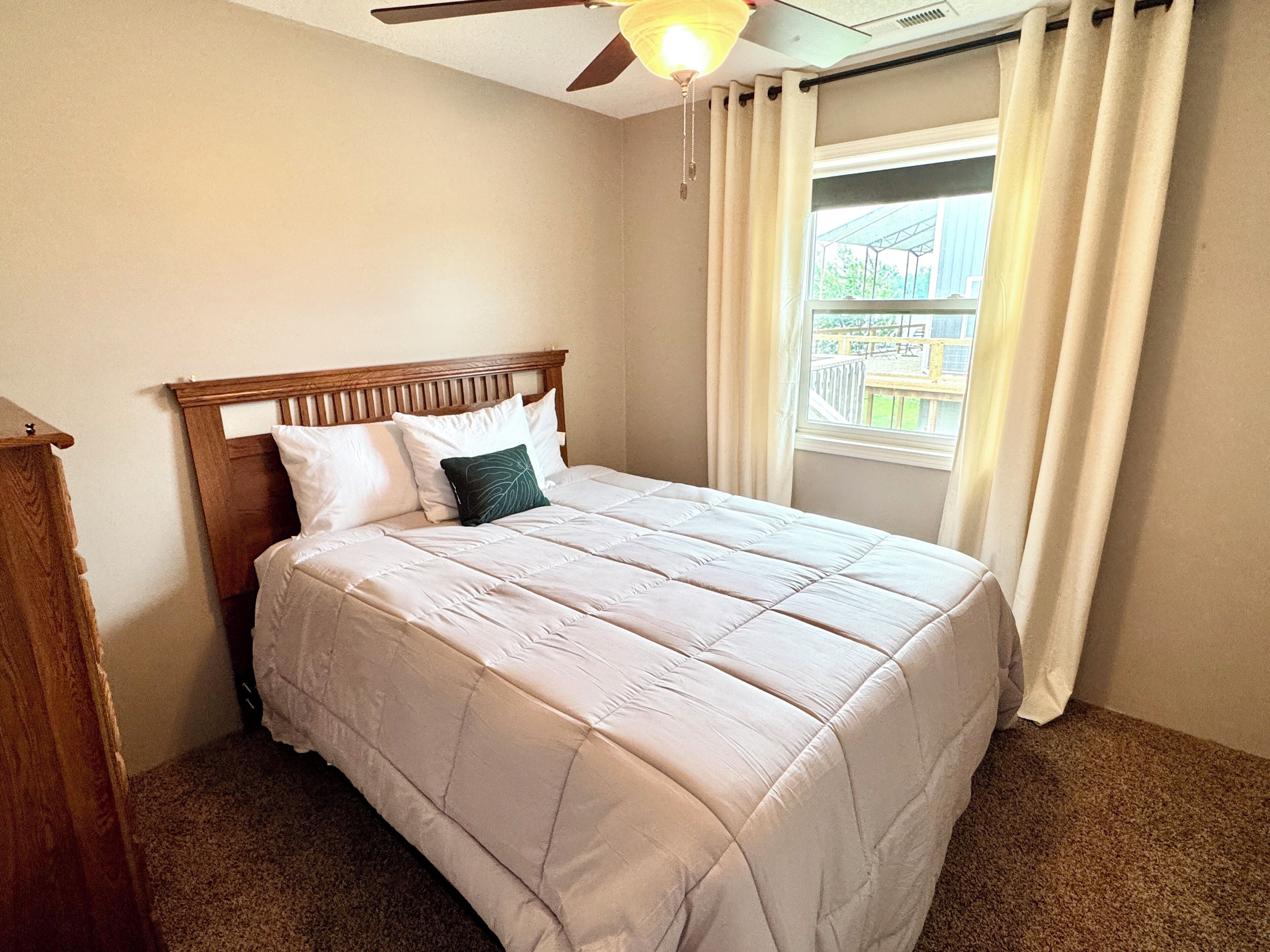 A bedroom with a bed, a ceiling fan, a window with curtains, and a view of the outside.