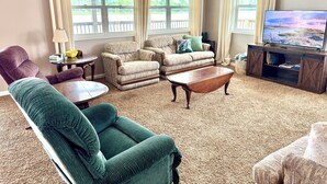 A living room with a sofa, armchairs, a coffee table, a TV, and large windows with curtains. - Lake Life Starts Here—w/ Dock, Kayaks & Relaxation (Craig)