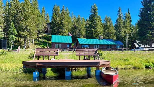 A wooden dock with benches and a boat, a cabin with a green roof, and a forested backdrop. - Harvey’s River Retreat 30 minutes from Yellowstone (Island Park)