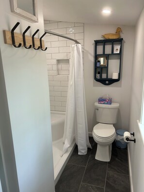 Un baño con una bañera blanca, un inodoro blanco, una cortina de ducha, una decoración de pato de madera y un estante pequeño con un espejo y una planta.